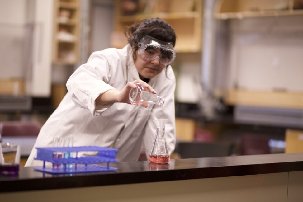 Indigenous student from the Ntekepmx Nations conducting an experiment by pouring liquid from one Erlenmeyer flask into another in a chemistry laboratory.