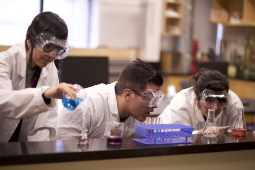 Three Indigenous students from the Haida and Ntekepmx Nations in a chemistry laboratory. One student is pouring liquid from one Erlenmeyer flask into another while the other two students are examining liquid in a different Erlenmeyer flask.