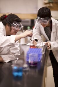 Two Indigenous students measuring liquids using Erlenmeyer flasks in a chemistry/biology laboratory.