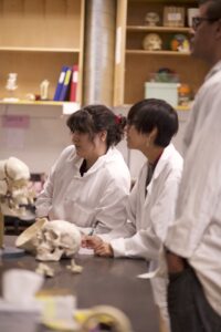 Three Indigenous students from Haida and Ntekepmx Nations gathered around a table with skull props on it in an anatomy laboratory. The students are paying attention to someone off camera.