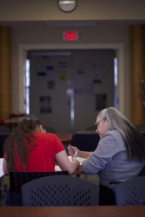 Two Indigenous students from the St'at'imc/Nlaka'pamux and Fort McKay First Nations sitting and studying in a campus common space with a clock above them.