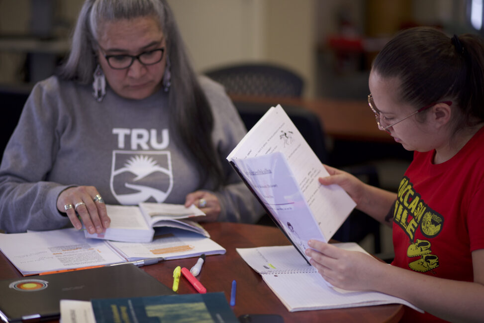 Two Indigenous students from the St'at'imc/Nlaka'pamux and Fort McKay First Nations studying and reading books at a table in a common space on campus.