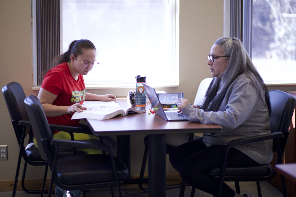 Two indigenous students from the Stat'imc/Nlakapamux and Fort McKay First Nations taking some notes and studying at a common space on campus. the student on the right is suing a computer and the one on the left is reading a book.