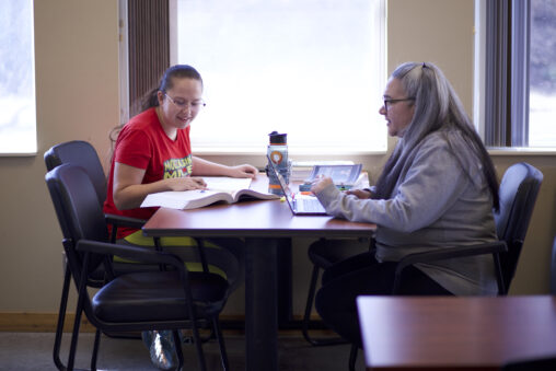 Two Indigenous students from the Stat'imc/Nlakapamux Fort McKay First Nations reading a physical book and a digital textbook in a campus common space.