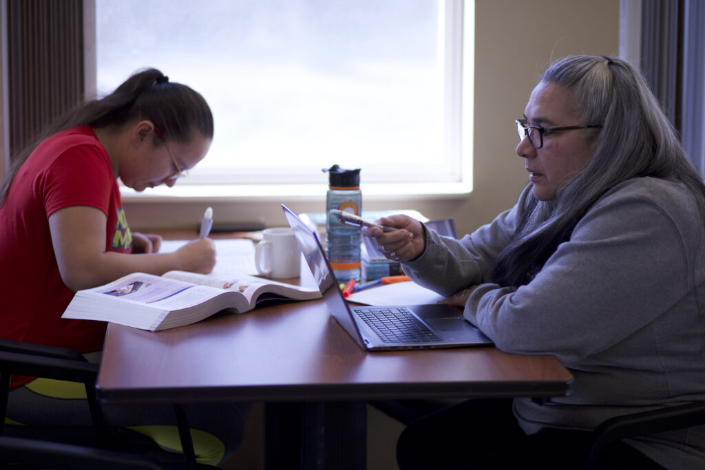 Two indigenous students from the Stat'imc/Nlakapamux Fort McKay First Nations studying at a common space on campus. The student on the right is using a computer and the on eon the left is taking some notes.