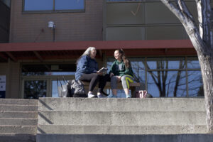 Two Indigenous students from the St'at'imc/Nlaka'pamux and Fort McKay First Nations sitting on an outdoor bench in front of a campus building.