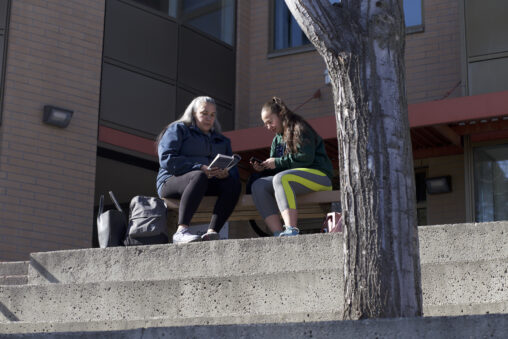 Two indigenous students from the St'at'imc/Nlaka'pamux and Fort McKay First Nations sitting on outdoor bench in front of a campus building.