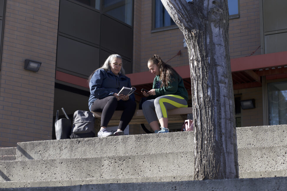 Two indigenous students from the St'at'imc/Nlaka'pamux and Fort McKay First Nations sitting on outdoor bench in front of a campus building.