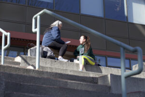 Two Indigenous students from the St'at'imc/Nlaka'pamux and Fort McKay First Nations sitting outdoors at the top of concrete stairs in front of a campus building.