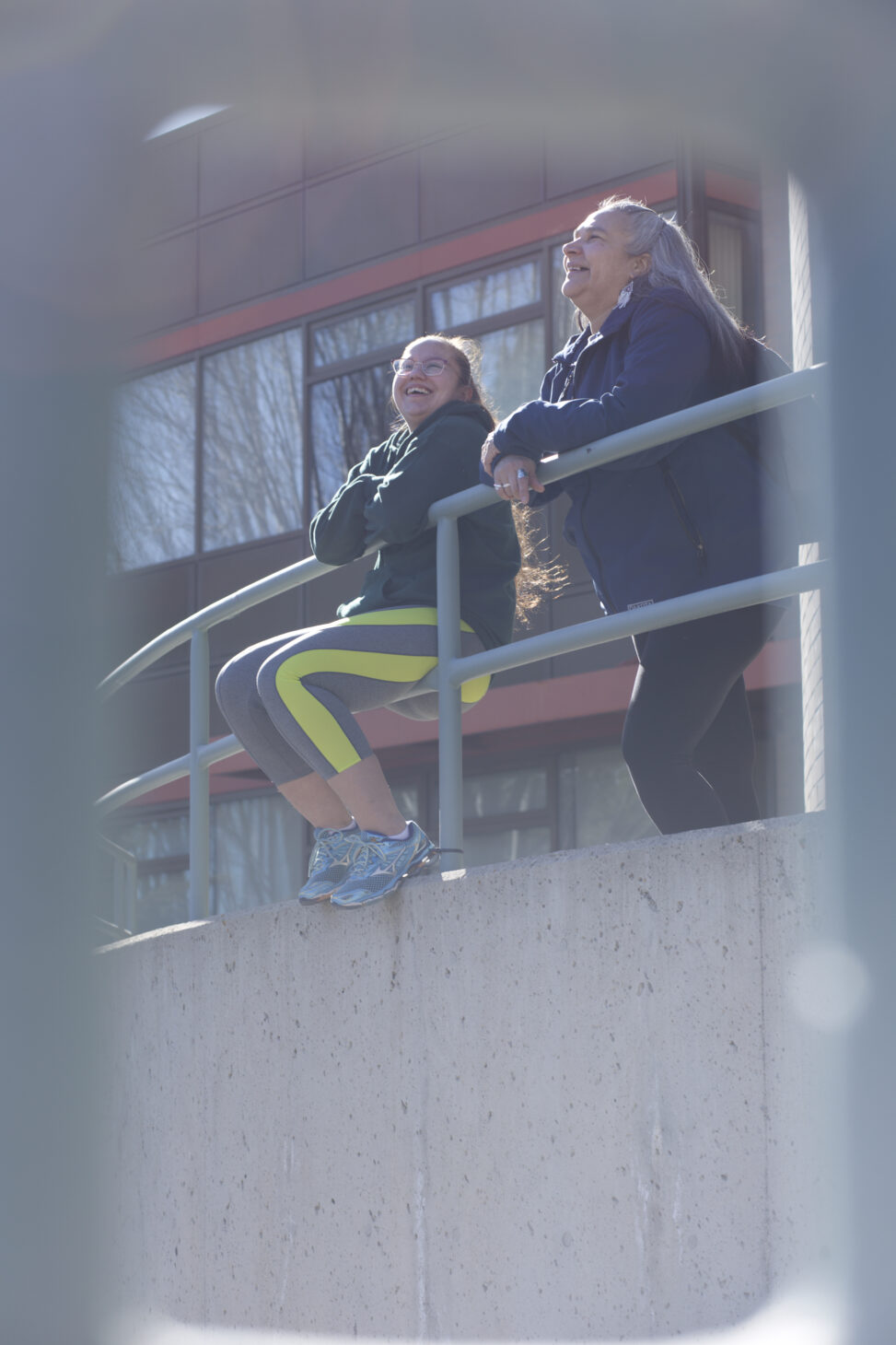 Two indigenous students Stat'imc/Nlaka'pamux and Fort McKay First Nations laughing in front of a campus building outdoors.