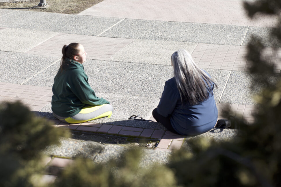 Two Indigenous students from the St'at'imc/Nlaka'pamux and Fort McKay First Nations sitting outdoors on concrete steps on a campus.