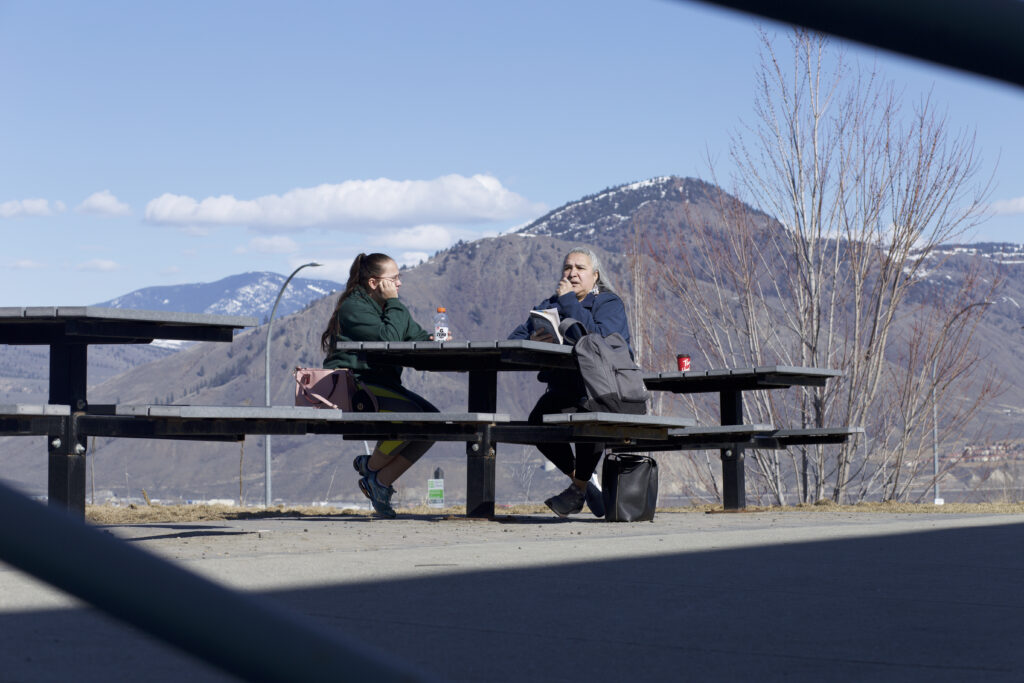 Two Indigenous students from the St'at'imc/Nlaka'pamux and Fort McKay First Nations sitting at an outdoor table with their backpacks and a book. Mountains in Kamloops are in the background.