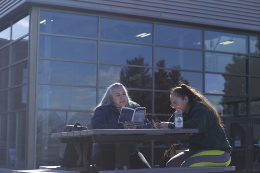 Two Indigenous students from the St'at'imc/Nlaka'pamux and Fort McKay First Nations sitting at an outdoor table. One of the students is reading a book while the other one is looking at their phone.